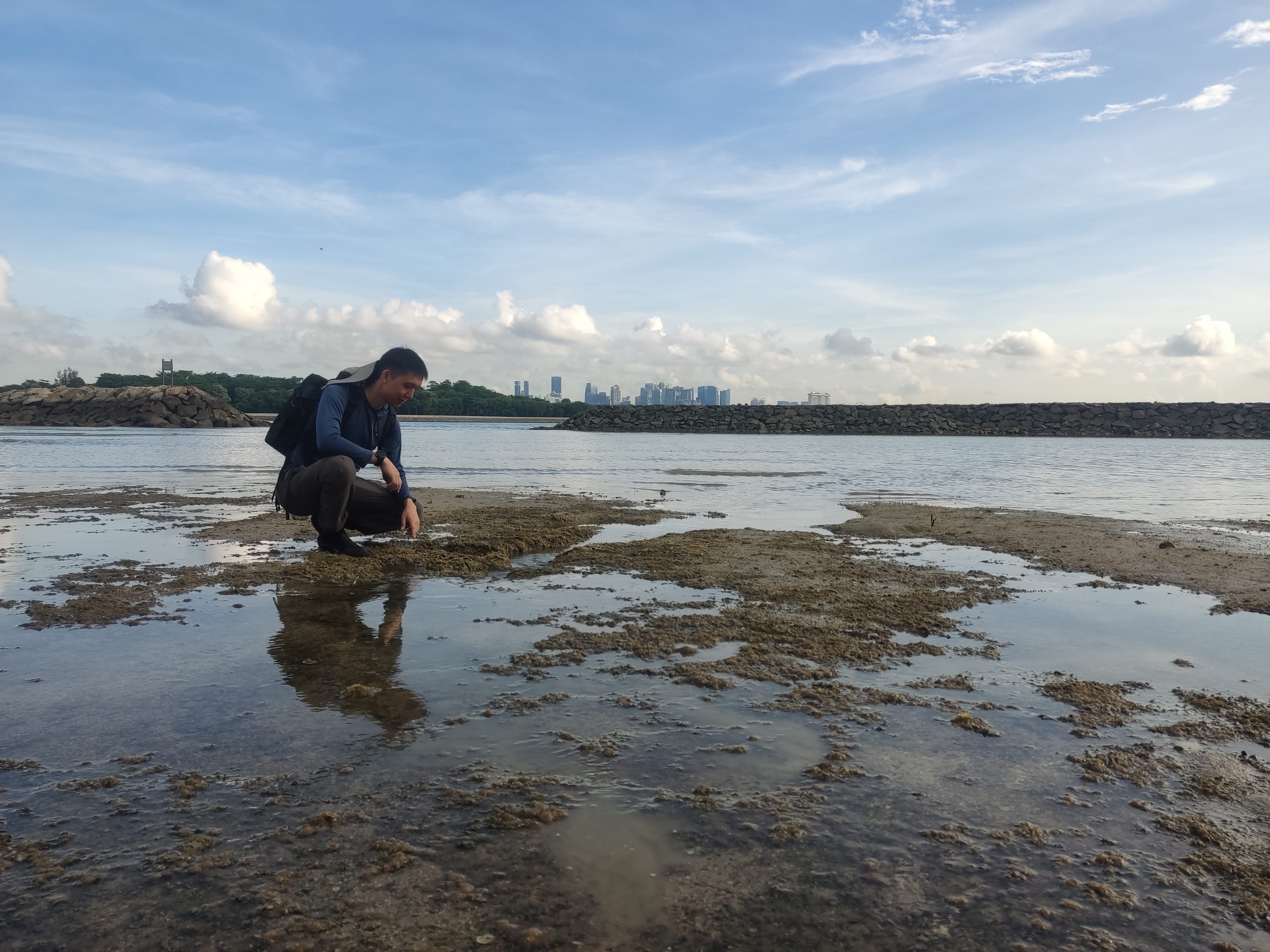 A person in squatting position on a rocky beach with the reflection on the water with a city skyline in the background under a blue sky.